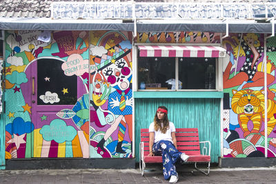 Full length portrait of woman sitting outside store with graffiti on bench