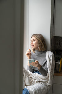 Smiling woman looking away while sitting by window with smart phone