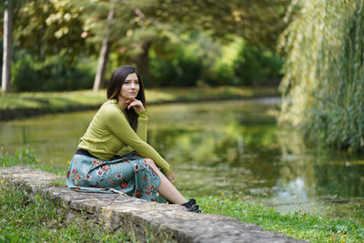 Full length of young woman sitting on grass by lake
