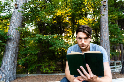 Young man reading book while sitting against trees at park