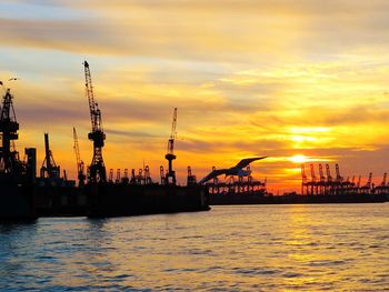 Silhouette cranes at harbor against sky during sunset