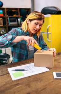 Blonde woman with cutter unpacking order received by courier with merchandise to sell on line