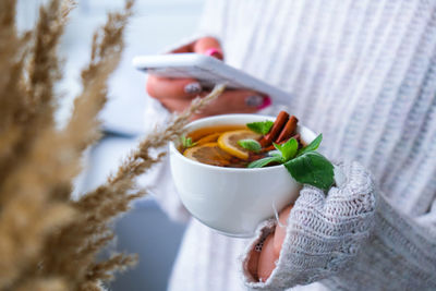 Female hands on sweater using phone and drinking tea with lemon, cinnamon sticks and mint leaves. 