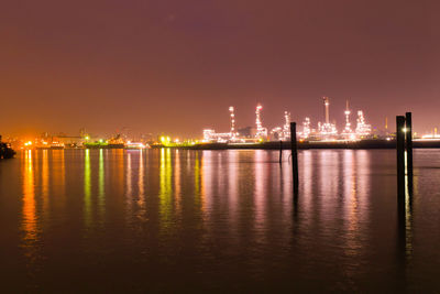 Illuminated factory by sea against sky at night