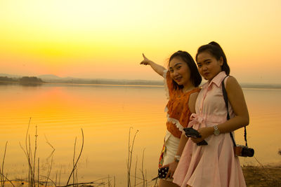 Portrait of smiling woman pointing towards lake while with friend on field during sunset