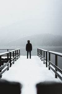 Rear view of man standing on snow covered pier by lake