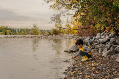 Boy crouching at lake against sky during autumn