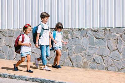 Schoolboy with backpack speaking with female friends while strolling on tiled pavement against stone wall in sunlight