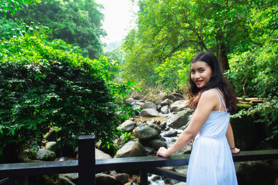 Portrait of smiling young woman standing by railing against trees