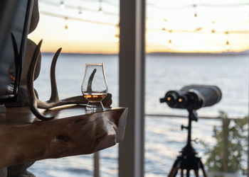 Close-up of drink in glass on table by window against sea during sunset