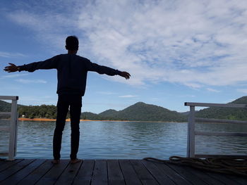 Rear view of man with arms outstretched standing on pier over lake against sky