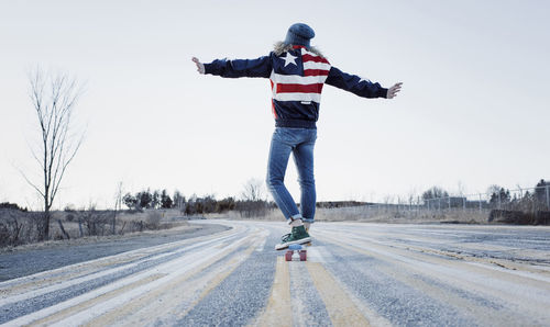 Rear view of carefree woman with arms outstretched skateboarding on road against clear sky during winter