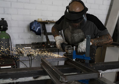 Side view of serious male welder using electric grinder and cutting metal detail in grungy workshop