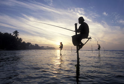 Silhouette fishermen stilt fishing in sea during sunset