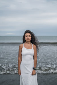 Portrait of young woman standing at beach against sky