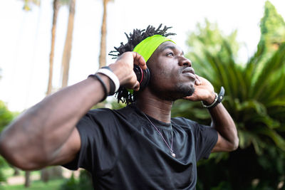 Young african american male athlete in sportswear listening to music with his headphones keeping hands on the headphones and looking away while standing in green tropical park