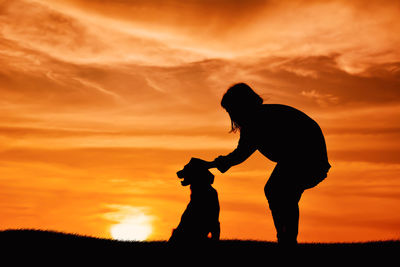 Silhouette girl petting dog while standing on field against sky during sunset