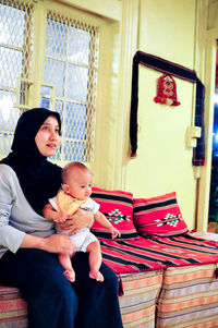 Mother holding son while sitting on bed at home