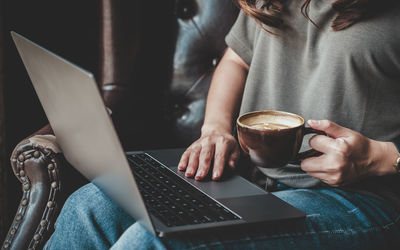 Midsection of businesswoman with coffee using laptop while sitting on chair in office