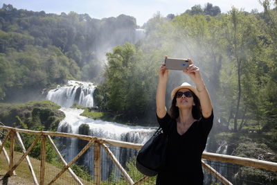 Smiling woman taking selfie while standing by railing with waterfall in background