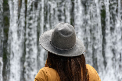 Rear view of woman wearing warm clothes and hat, standing in front of waterfall.