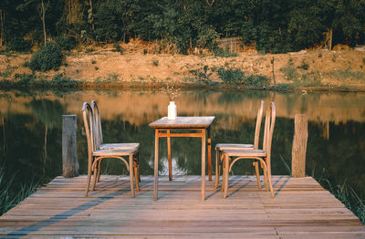 Empty chairs and table on pier over lake against trees