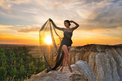 Young woman standing on rock against sky during sunset