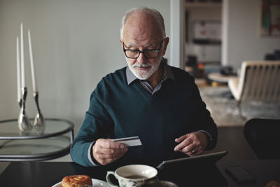 Wrinkled man holding credit card by digital tablet while sitting at dining table in living room
