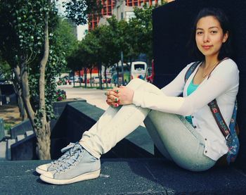 Full length portrait of young woman relaxing on retaining wall in city