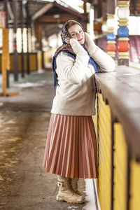 A slavic woman in a national colored scarf,a fur coat and  mittens on the porch of a wooden house