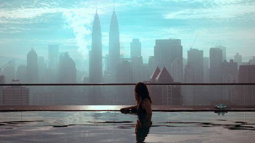 Rear view of woman standing by swimming pool against buildings in city