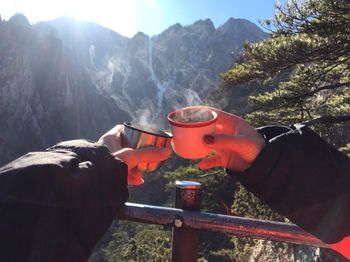 Cropped hands of couple toasting cups against mountain on sunny day