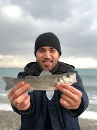 Portrait of smiling young man holding fish while standing at beach against cloudy sky