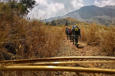 Rear view of people walking on dirt road amidst dry grass against mountains