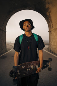 Portrait of young man holding skateboard while standing in tunnel