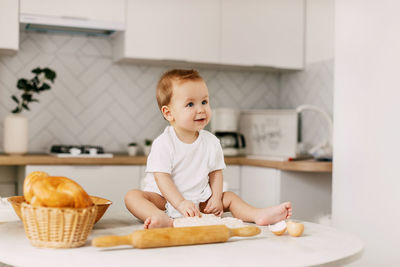 A little boy is sitting on the kitchen table with a rolling pin and a baking dough. 