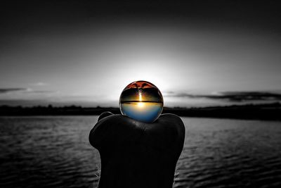 Cropped hand holding glass ball with upside down reflection on sea against sky
