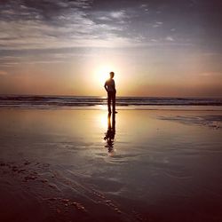 Silhouette teenage boy standing at beach against sky during sunset