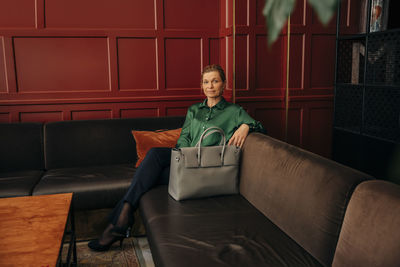 Full length portrait of mature businesswoman with bag sitting on sofa in hotel lounge