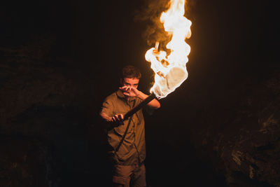 Young male speleologist with flaming torch standing in dark narrow rocky cave while exploring subterranean environment