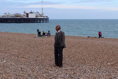 Side view of old man in suit on pebble beach with pier in background