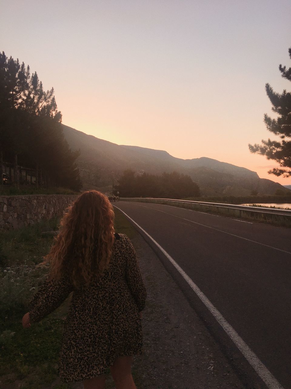 REAR VIEW OF WOMAN STANDING BY ROAD AGAINST SKY DURING SUNSET
