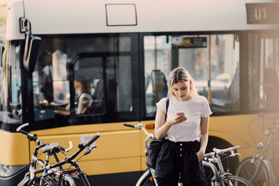 Young woman using smart phone while standing by bicycles against bus in city