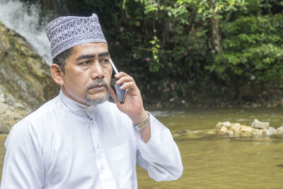 Thoughtful man talking on phone while sitting against waterfall