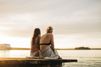 Rear view of female friends looking at lake while sitting on jetty during sunset