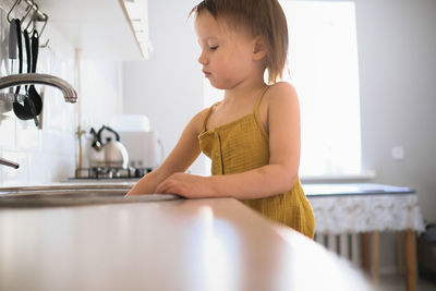 European child of four years old in yellow jumpsuit washes his hands in real bright kitchen