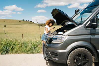 Young ethnic female with afro hair calling to repair service on smartphone while standing near camper van with open hood on countryside road in summer day