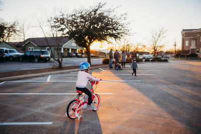 Young girl riding bike towards family in parking lot at sunset
