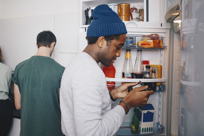 Side view of man looking at food package standing by refrigerator in kitchen