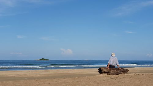 Rear view of man sitting on log at beach against sky